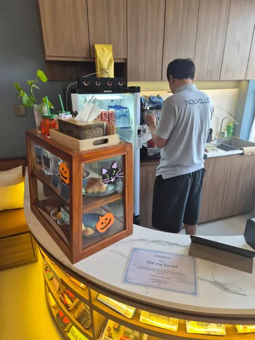 Nouvelle's coffee bar: a barista in a Nouvelle shirt at the espresso machine, a timber pastry cabinet with a croissant and madeleines, and books lit beneath the marble counter