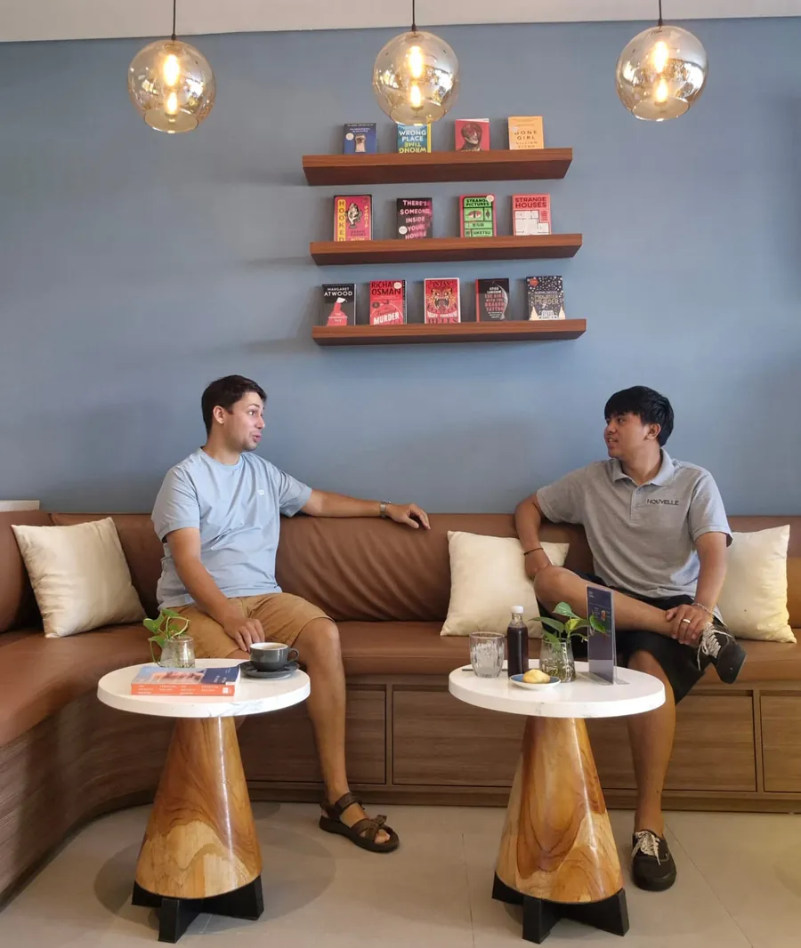 Two people talking over coffee on Nouvelle's caramel leather banquette, under amber globe lamps and shelves of face-out novels on the blue wall