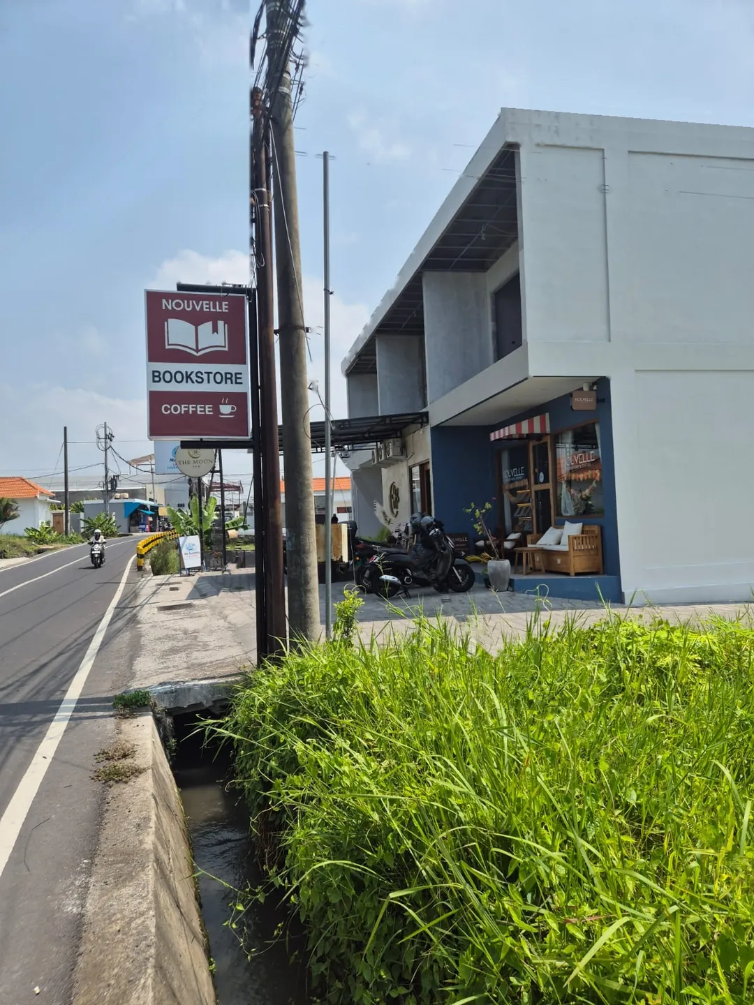The maroon Nouvelle Bookstore Coffee sign beside Jalan Pantai Seseh, with the shop's blue frontage and striped awning behind it